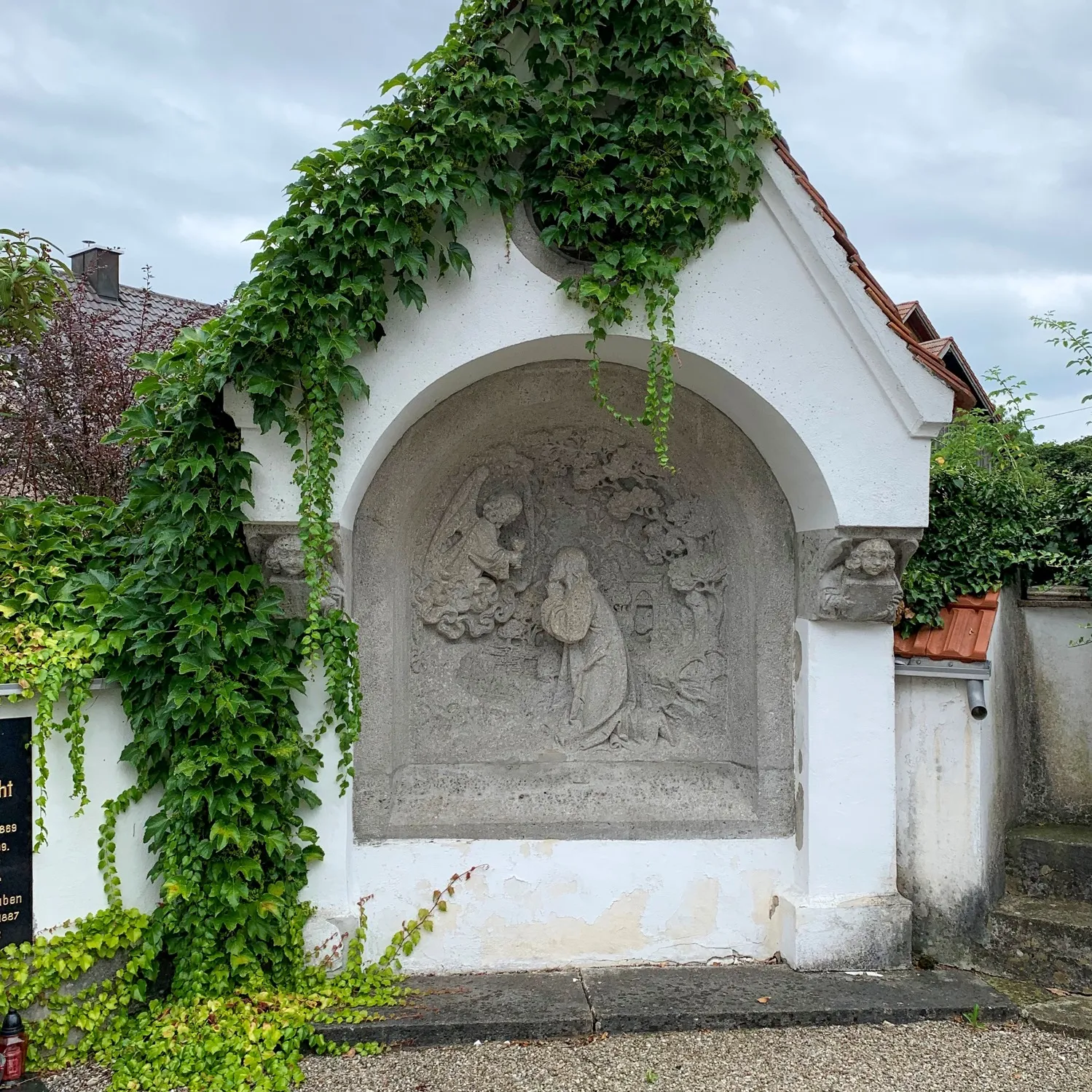 Ein weiß gekalkter Bildstock mit steinernem Relief, dicht von Efeu überwachsen.