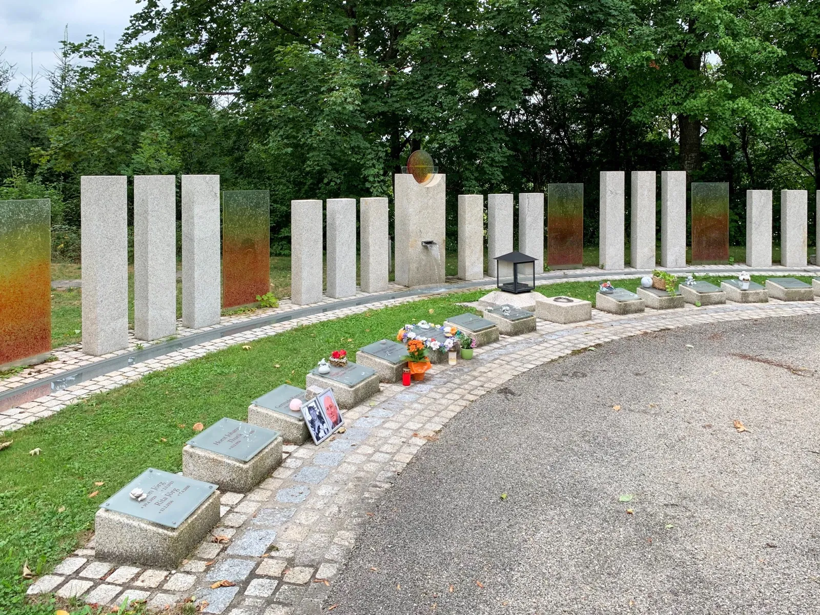 Eine geschwungene Reihe heller Stelen auf einer Rasenfläche, davor niedrige Grabplatten mit Blumenschmuck und Grablaternen.