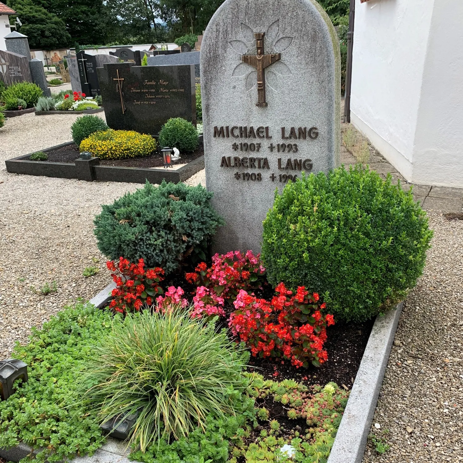 Ein Erdgrab mit steinernem Kreuz, davor rote Blumen, kleine Buchsbaumkugeln und dicht bepflanzte Bodendecker.