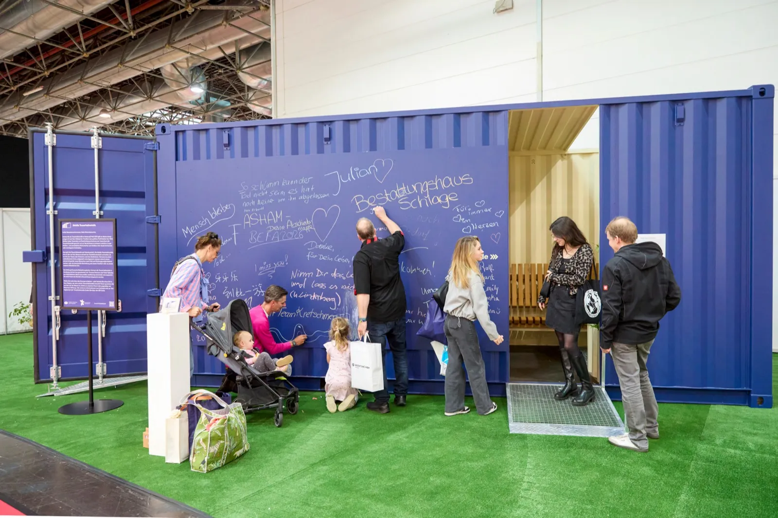 Ein blau lackierter Überseecontainer als mobile Trauerhaltestelle auf einer Messe; Besucherinnen und Besucher schreiben auf die beschriftete Wand.