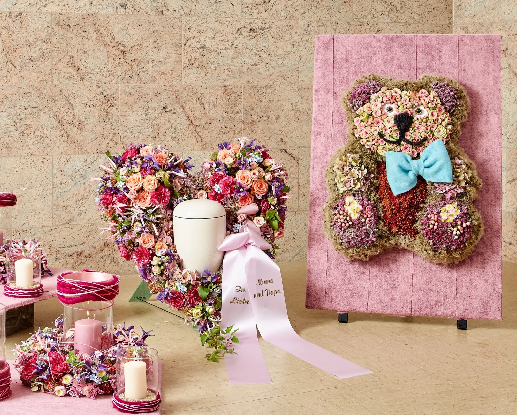 Blumenschmuck in Rosa- und Violetttönen: ein Herz aus Blüten, eine helle Urne mit Schleife und ein aus Blumen gesteckter Teddybär auf einer rosa Tafel.