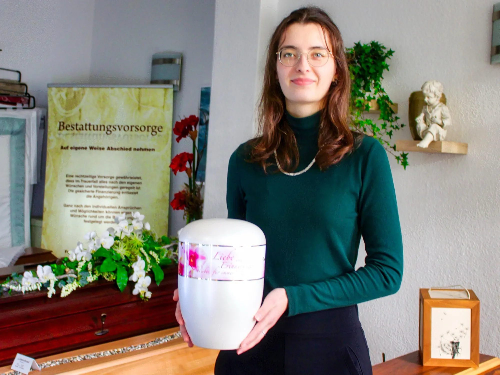 Eine junge Frau mit Brille im dunkelgrünen Rollkragenpullover hält eine helle Urne in beiden Händen, dahinter ein Ausstellungsraum mit Blumen und einem Plakat zur Bestattungsvorsorge.