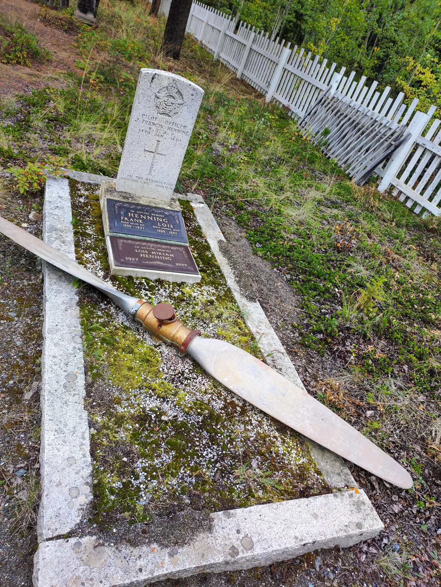 Ein helles Grabmal mit eingemeißeltem Kreuz, davor liegt ein hölzerner Flugzeugpropeller auf der steinernen Einfassung.