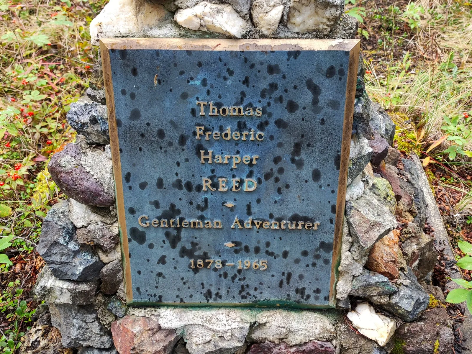 Eine verwitterte Bronzetafel für Thomas Frederic Harper Reed, eingefasst in einen Hügel aus bunten Steinen.