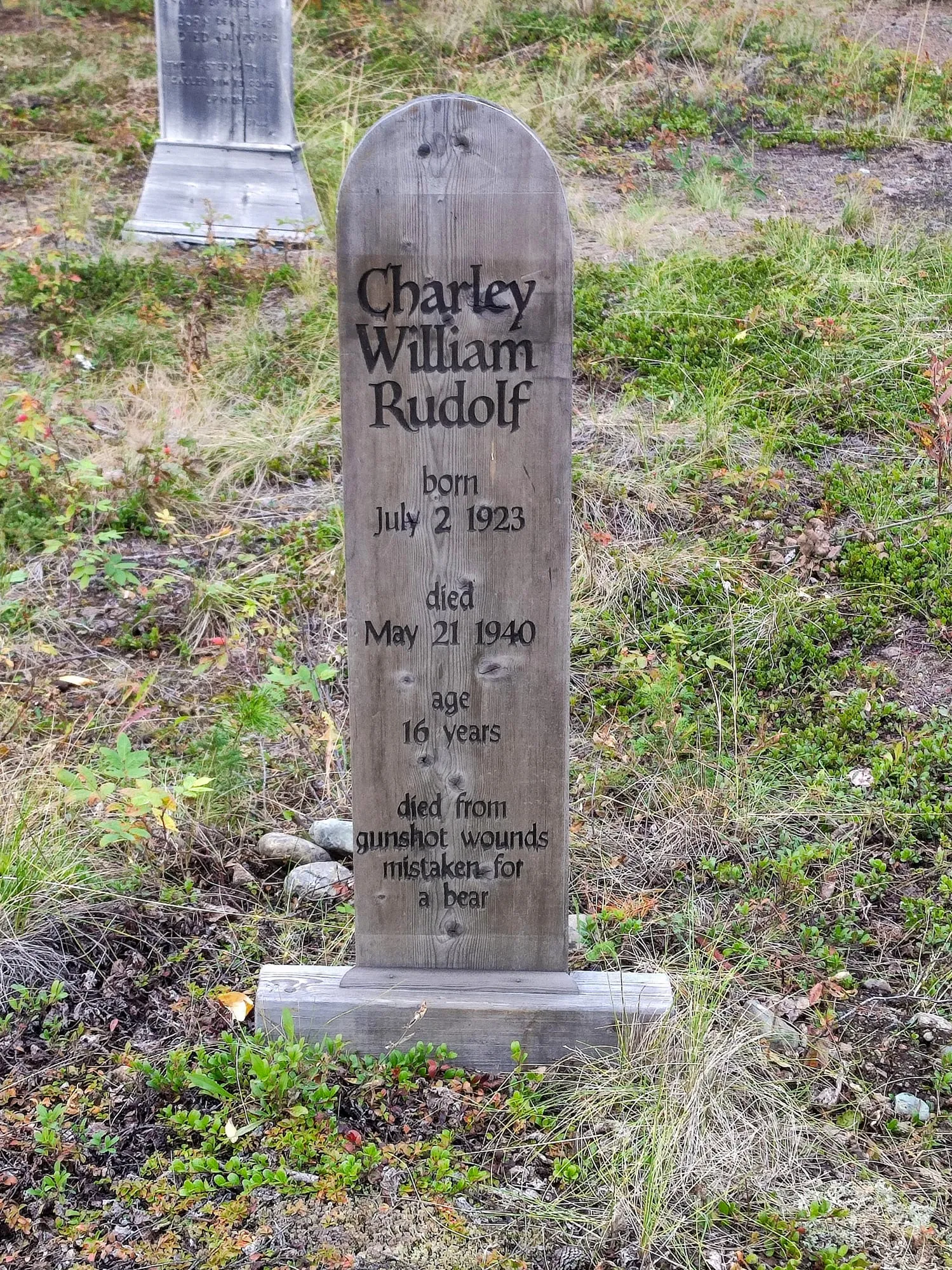 Ein hölzerner Grabstein auf einer Wiese, in den Name und Lebensdaten von Charley William Rudolf eingeschnitten sind.